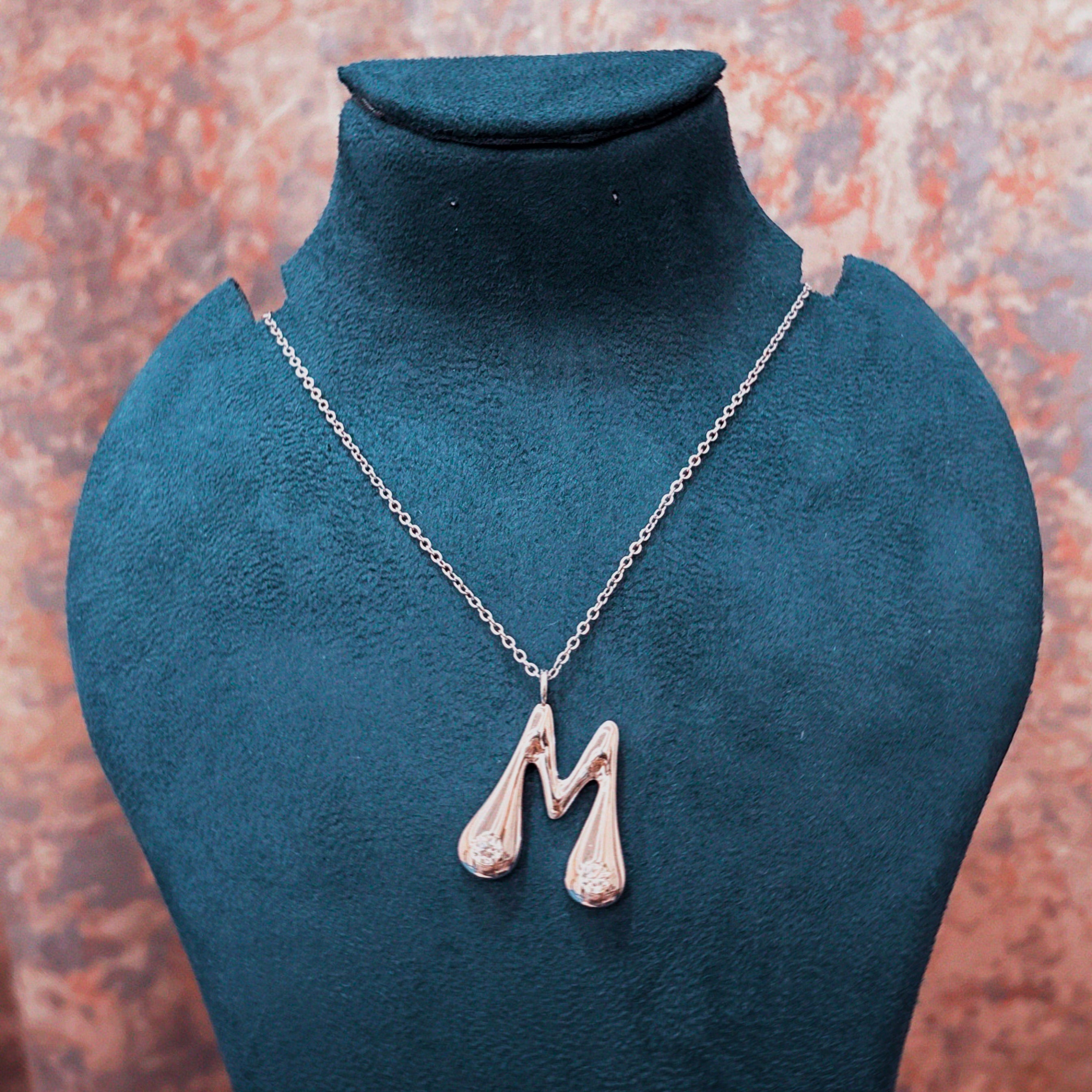 A silver-tone initial pendant necklace spelling 'M' with small clear stones, displayed on a dark teal jewelry bust against a textured orange-brown background.
